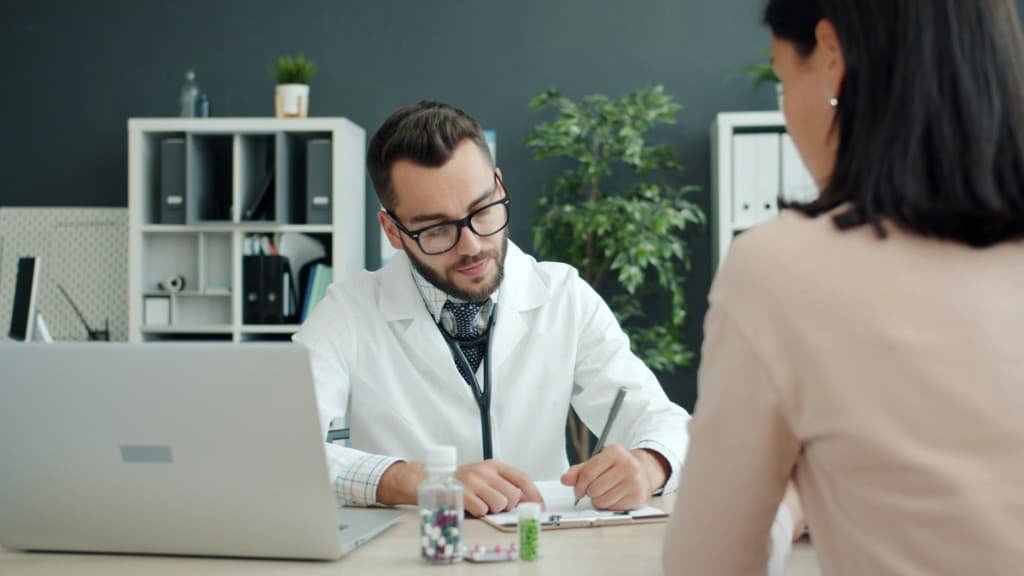 A physician in a white coat taking notes during a patient consultation at a clinical desk.